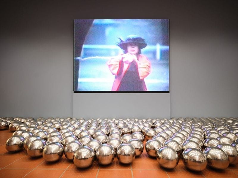 A photograph of stainless steel balls on a red brick floor before a monitor playing a video of a woman in an orange dress and black hat mounted on a plain gray wall
