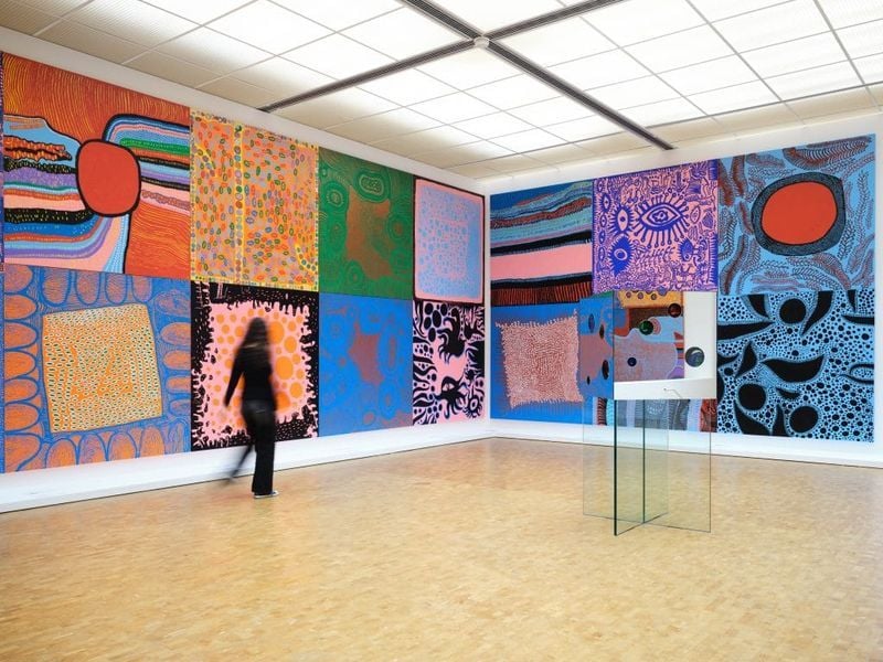 벽에는 다채로운 야요이 쿠사마의 추상 작품들이 벽을 장식하고 있는 완전히 천창이 들어간 미술관을 탐험하는 손님의 사진입니다