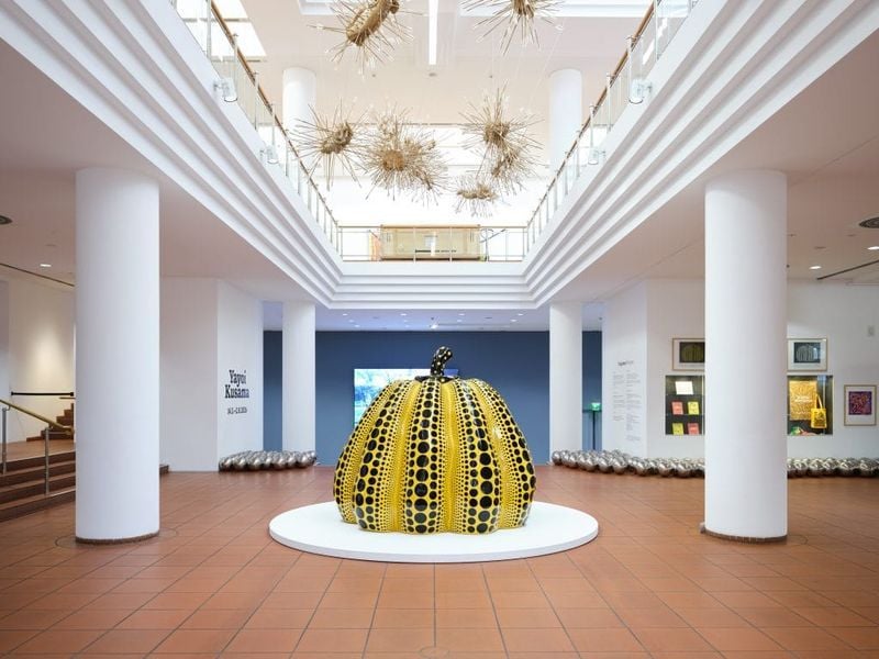 A photograph of a large iconic yellow pumpkin sculpture by Yayoi Kusama in the center of a white-walled, two tiered gallery hall, surrounded by columns