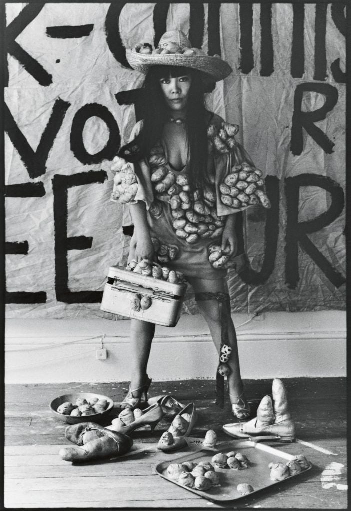 A black and white photogaph of a young Yayoi Kusama wearing a sculpturally adorned cowboy hat and dress surrounded my small, vaugely phallic sculptures standing before a mural with text in her studio