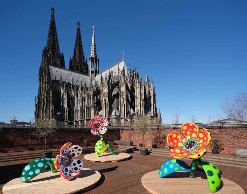 맑고 푸른 하늘 아래 갈색 지붕 테라스 위에 다채로운 꽃 조각들이 놓여 있고, 배경에는 위엄 있는 고딕 양식의 대성당이 우뚝 서 있는 사진입니다