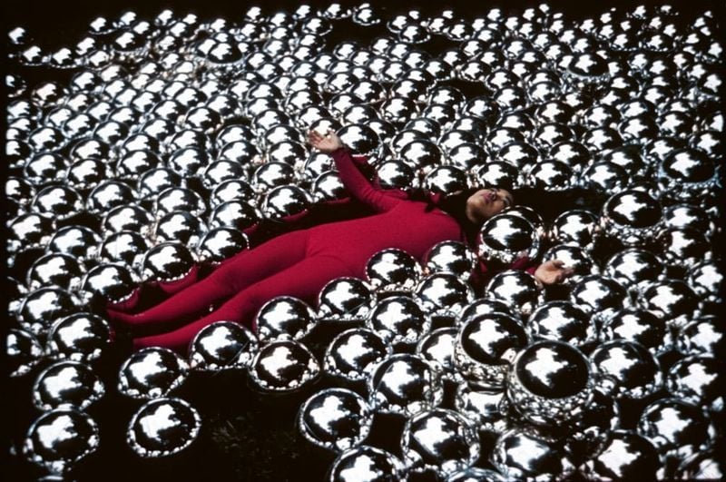 빨간 바디수트를 입은 쿠사마 야요이가 반사되는 은빛 구체 사이에 누워 있어 몰입감 있는 거울 아트 설치 환경을 만들어낸다